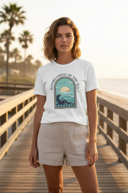 White t-shirt with a graphic design of a woman on a beach and text 'Daydreaming of the Sea' on a white background.